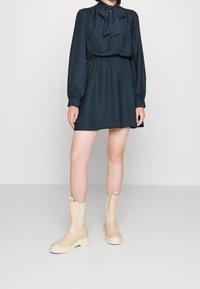 Tummanvihreä, teksturoitu mekko, jossa on rusetti kauluksessa ja pitkät hihat, yhdistettynä beigen sävyisiin nilkkureihin, joissa on joustavat sivuosat ja paksut pohjat.