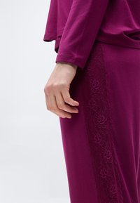 Χέρι που ακουμπάει στο πλάι, φοράει σκούρο ματζέντα σύνολο με μακρυμάνικη μπλούζα και παντελόνι που έχει λεπτομέρειες με λουλουδάτη δαντέλα στο πλάι.