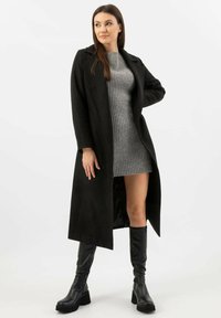 Černý dlouhý kabát se strukturovaným designem, nošený přes šedé žebrované šatové šaty a vysoké černé kožené boty, zdůrazňující vrstvený styl.