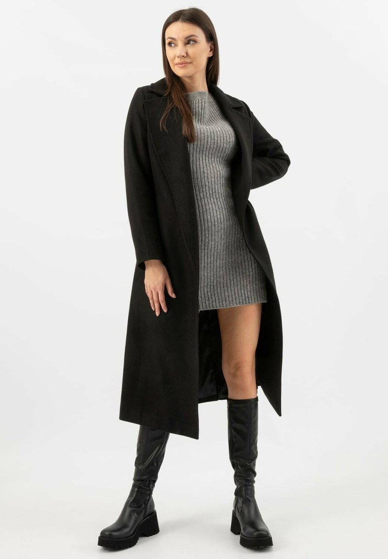 Černý dlouhý kabát se strukturovaným designem, nošený přes šedé žebrované šatové šaty a vysoké černé kožené boty, zdůrazňující vrstvený styl.