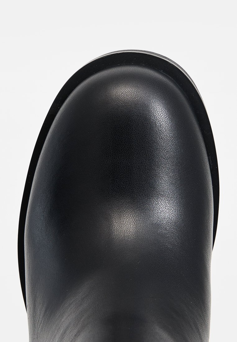 Bottine en cuir noir avec un bout rond et une texture lisse, présentant un motif de grain subtil et une silhouette élégante.