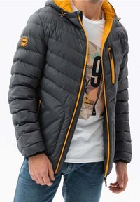 Chaqueta acolchada en gris oscuro con acentos naranjas, que incluye una capucha, bolsillo en el pecho con cremallera y bolsillos laterales. Diseño texturizado y acolchado.