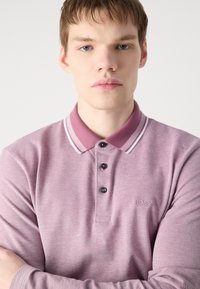 Ljubičasta polo majica dugih rukava s jedinstvenim uzorkom, s kontrastnim ovratnikom s bijelim prugama, tri crna gumba i logotipom.