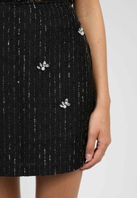 Mini-jupe en tweed noir avec des rayures verticales en fils argentés et des ornements décoratifs en forme de fleurs avec des perles et des cristaux.