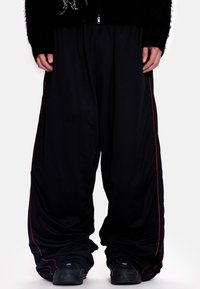 Juodos tinklelio plataus kirpimo kelnės su raudonomis šoninėmis juostelėmis. Turi laisvą siluetą ir tekstūruotą paviršių, derinamos su juodais sportbačiais.