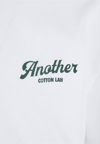 Weißer Stoff mit dunkelgrünem Text, der "Another" in großer Kursivschrift und "COTTON LAB" in kleineren Großbuchstaben darunter zeigt.