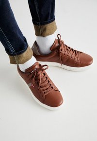 Bruna lädersneakers med rund tå, vit gummisula och bruna snören. Har varumärkeslogotyp på tungan och sidan, tillsammans med mörk denimjeans.