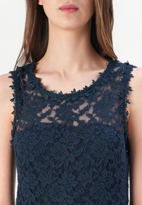Námořnická modrá krajková šaty s květinovým vzorem a strukturovaným povrchem. Má scalloped výstřih a ramenní detaily s květinovými motivy.