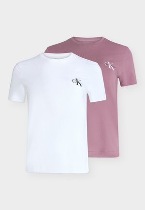 Jeune femme debout avec une main dans la poche, portant un t-shirt rose clair et un jean ample bleu foncé sur un fond gris uni.