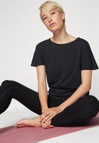 Maglietta nera a maniche corte con scollatura tonda abbinata a leggings neri aderenti, seduta su un tappetino rosa testurizzato, con uno sfondo neutro.