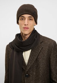 Brown knitted beanie and matching scarf worn over a textured brown coat. The scarf drapes softly, complementing the relaxed fit of the outfit.