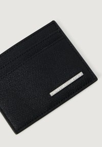 Crni kožni novčanik s teksturom s dva utora za kartice i srebreni metalni Armani Exchange logo na bijeloj pozadini.