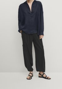 Laivastonsininen pellavainen pitkähihainen paita, jossa on rento istuvuus ja avoin kaulus, yhdistettynä löysiin mustiin cargo-housuihin ja mustiin remmikkäisiin sandaaleihin.