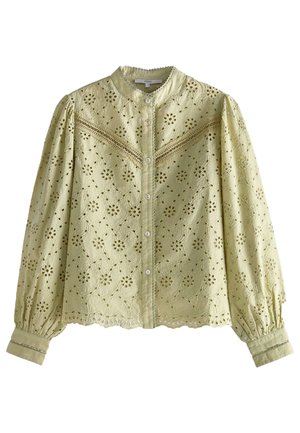 Femme portant un chemisier vert clair ajouré, un jean slim bleu et des escarpins slingback en peau de serpent, debout sur un fond rose pâle.