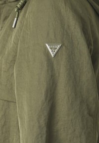 Giacca verde oliva, texturizzata, con cappuccio e un logo triangolare argento sulla manica sinistra. Presenta un cordoncino al collo.