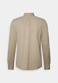 Beige långärmad skjorta med knäppningar i ärmsluten, krage och en mitt bakveck, visad bakifrån mot en vit bakgrund.