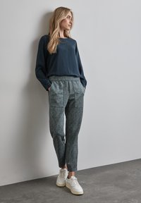 Navy, lange mouwen top met een gladde textuur, gecombineerd met gemusterde, taps toelopende broeken in teal en crèmekleurig. Model draagt witte sneakers.