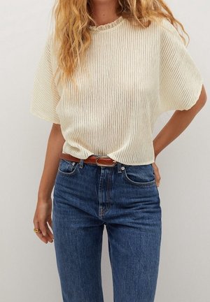 Mujer con una camiseta corta de manga corta color crema con textura acanalada, metida dentro de unos vaqueros azules de talle alto con un cinturón de cuero marrón, de pie con una mano en la cadera.