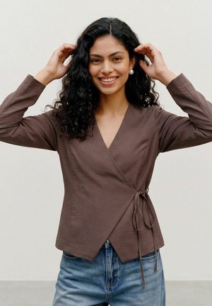 Femme aux cheveux noirs bouclés portant un haut cache-cœur marron et un jean bleu ajuste ses cheveux en souriant sur un fond clair uni.