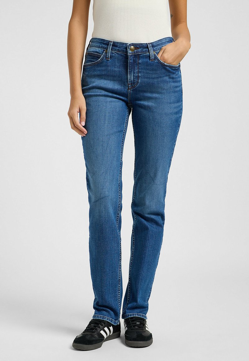 Vaqueros de mezclilla azul con un corte ajustado, diseño de cinco bolsillos y un ligero desgaste. Combinados con zapatillas negras con franjas blancas.