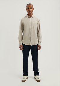 Helle beige Langarmbluse mit Knopfleiste und einer Brusttasche, kombiniert mit navyblauen Slim-Fit-Hosen und weißen Sneakers mit braunen Sohlen.