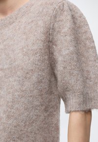 Gros plan sur une personne portant un pull tricoté doux, duveteux, de couleur marron clair avec des manches courtes légèrement bouffantes.