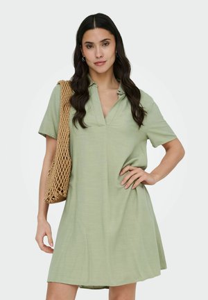 Ženska v kratki obleki zelene barve žajblja z ovratnikom, ki nosi tkan bež torbo čez ramo, pozira z eno roko na boku.