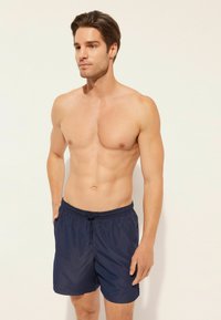 Mörkblå badshorts med resårmidja och dragsko. Slät, lättviktsmaterial, knälång skärning, utan mönster eller dekorationer.