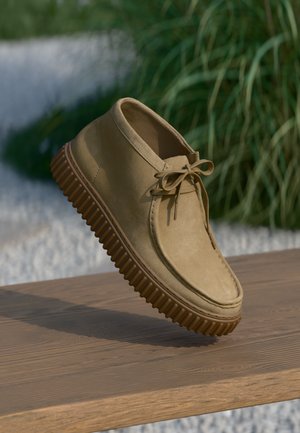 Stivaletti alla caviglia in suede beige con punta arrotondata, chiusura con lacci e suola in gomma marrone texture. Sullo sfondo si vedono erba e ghiaia.