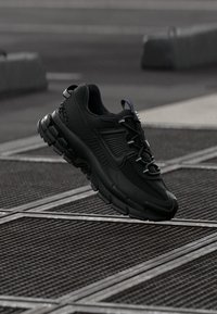 Čierne bežecké tenisky Nike s textúrovanou podrážkou a šnúrkami, suspendované nad kovovou mriežkovou dlažbou v mestskom vonkajšom prostredí.