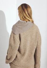 Beige stickad tröja med en stor, mjuk taupe-färgad krage. Tyget ser fluffigt ut, med en avslappnad passform och sänkta axelsömmar.