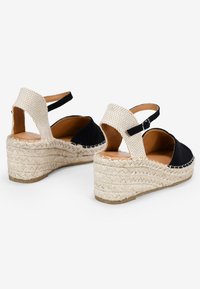 Alpargatas de cuña en lona negra con acentos trenzados de color beige y una correa. Presentan una suela de cuerda texturizada y una plantilla de cuero natural.