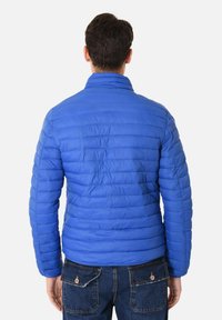 Giacca blu imbottita con colletto rialzato, caratterizzata da un design a cuciture orizzontali e una texture liscia. Indossata sopra jeans in denim scuri.