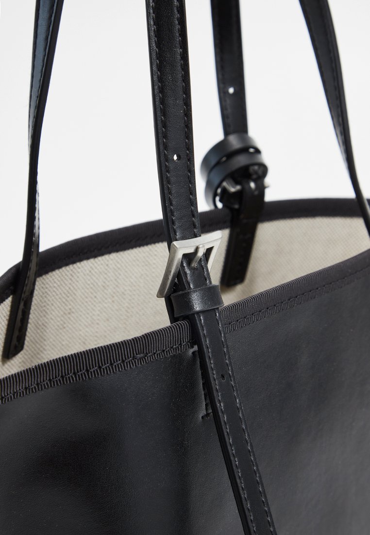 Schwarze Ledertasche mit glatter Oberfläche, ausgestattet mit einer silbernen Schnalle, verstellbaren Trägern und einem kontrastierenden beigen Innenfutter.