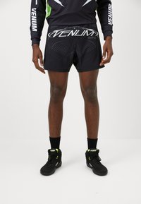 Schwarze Sportshorts mit enganliegendem Design, versehen mit einem silbernen Logo und geometrischem Muster. Kombiniert mit schwarzen Schuhen und schwarzen Socken.
