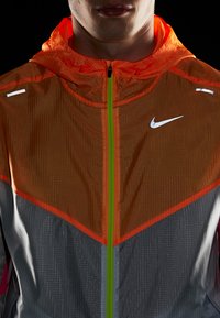 La chaqueta de Nike presenta un diseño en bloques de color naranja y gris, con un zipper verde, acentos reflectantes y una tela ligera y texturizada.