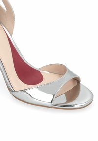 Sandale à talon haut argentée à bout ouvert avec des lanières croisées et une semelle intérieure rembourrée bordeaux sur fond blanc.