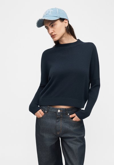 Γυναίκα που φοράει μπλε σκούρο πουλόβερ, σκούρο μπλε τζιν με τα χέρια στις τσέπες και ένα ανοιχτό μπλε καπέλο τζιν, κοιτώντας προς τα πλάγια μπροστά σε ένα απλό φόντο.