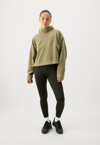 Hoodie verde oliva corto con tasca frontale e cappuccio con cordino, abbinato a leggings neri e sneakers nere. Dettaglio del logo bianco visibile.