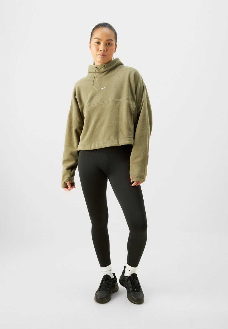 Olijfgroen cropped hoodie met een voorzak en een capuchon met trekkoord, gecombineerd met zwarte leggings en zwarte sneakers. Witte logo detail zichtbaar.
