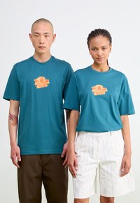 Teal cotton t-shirts with a central orange graphic. One model wears brown pants, the other white pinstriped shorts. Both feature short sleeves.