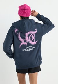 Marinegenser med et stort rosa grafisk design på ryggen, som inneholder tekst og en stilisert logo. Brukt med en lys rosa strikket lue.