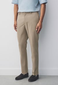 Pantalon beige texturé à coupe ajustée, avec une taille plate, des passants pour ceinture, et des poches latérales, associé à des mocassins en daim noir.