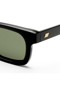Μαύρα ορθογώνια γυαλιά ηλίου με φακούς με πράσινη απόχρωση και χρυσό λογότυπο με τρεις γραμμές στο πλαϊνό μπράτσο, πάνω σε λευκό φόντο.