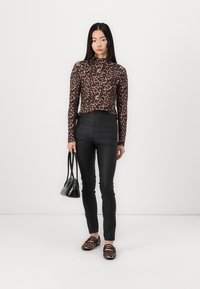 Bruine luipaardprint lange mouwtop, zwarte aansluitende broek en gemusterde ballerina's. Model houdt een kleine zwarte handtas vast tegen een witte achtergrond.