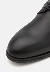 Zapato de cuero negro con una textura suave, puntera redondeada y detalles cosidos a lo largo de la suela, con un diseño minimalista.