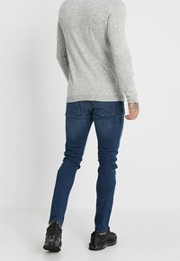 Camisa gris de manga larga, jeans ajustados azules y zapatillas negras con suela texturizada, vista desde atrás.