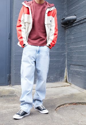 Personne portant une veste rouge et beige, un sweat-shirt bordeaux, un jean bleu clair et des baskets noires et blanches, debout contre un mur gris.