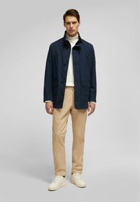 Marineblauwe lichtgewicht jacket, met knopen aan de voorkant, hoge kraag en twee voorzakken. Gekleed met beige broek en witte sneakers.