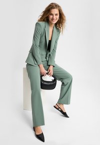 Groen op maat gemaakt pak met uitlopende broeken, gladde stof en scherpe revers. Model houdt een kleine zwarte handtas vast en draagt puntige zwarte slingback schoenen.